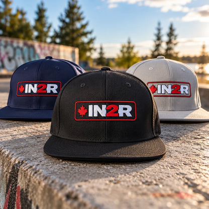 IN2R Canadian Edition snapback hat collection in black, navy blue and grey, featuring embroidered red maple leaf and red and white IN2R logos, displayed outdoors on concrete in warm sunlight.