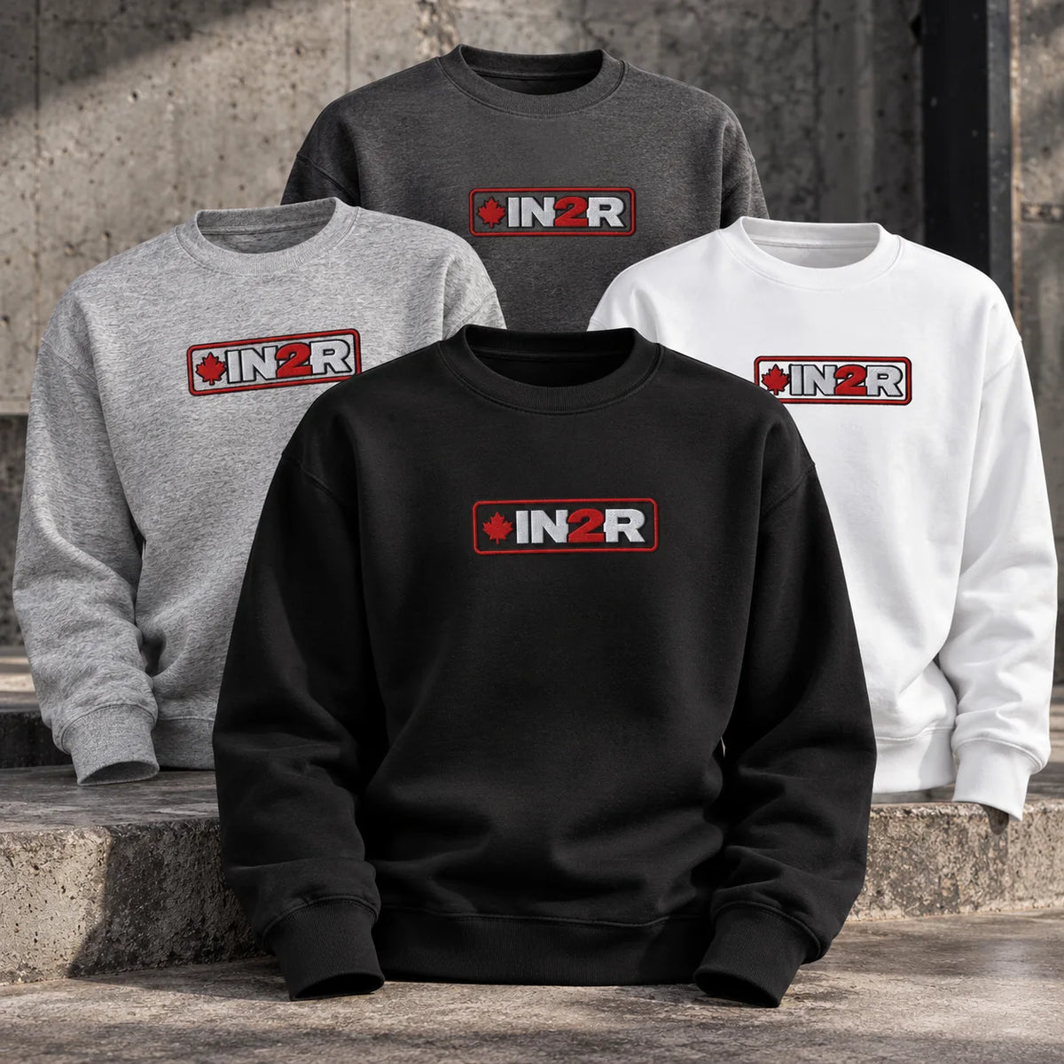 IN2R Canadian Edition crewneck sweatshirts in black, charcoal heather, carbon grey and white, featuring embroidered red maple leaf logos, displayed on concrete steps.