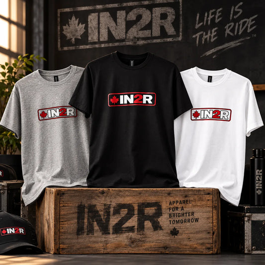 Three IN2R short-sleeve t-shirts displayed on a rustic wooden crate in a styled indoor setting. The shirts are shown in heather gray, black, and white, each featuring a red and white rectangular chest logo with a maple leaf and the text 'IN2R'.
