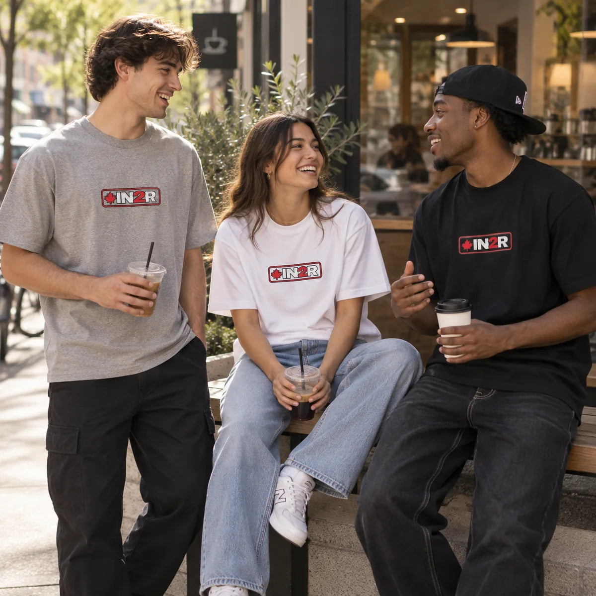 Three people standing and sitting outdoors in front of a storefront, wearing matching graphic T-shirts with a red, white, and black rectangular logo that reads 'IN2R'.