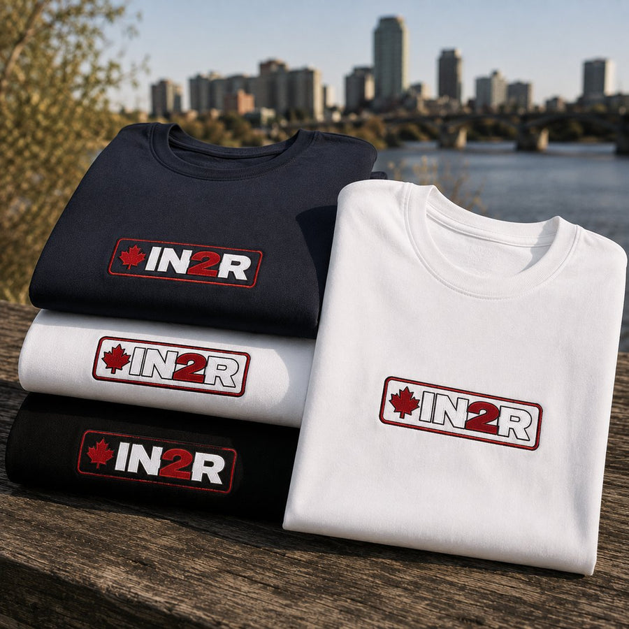 IN2R Canadian Edition black, white and navy tees arranged on a wooden riverfront table with embroidered maple leaf logos.