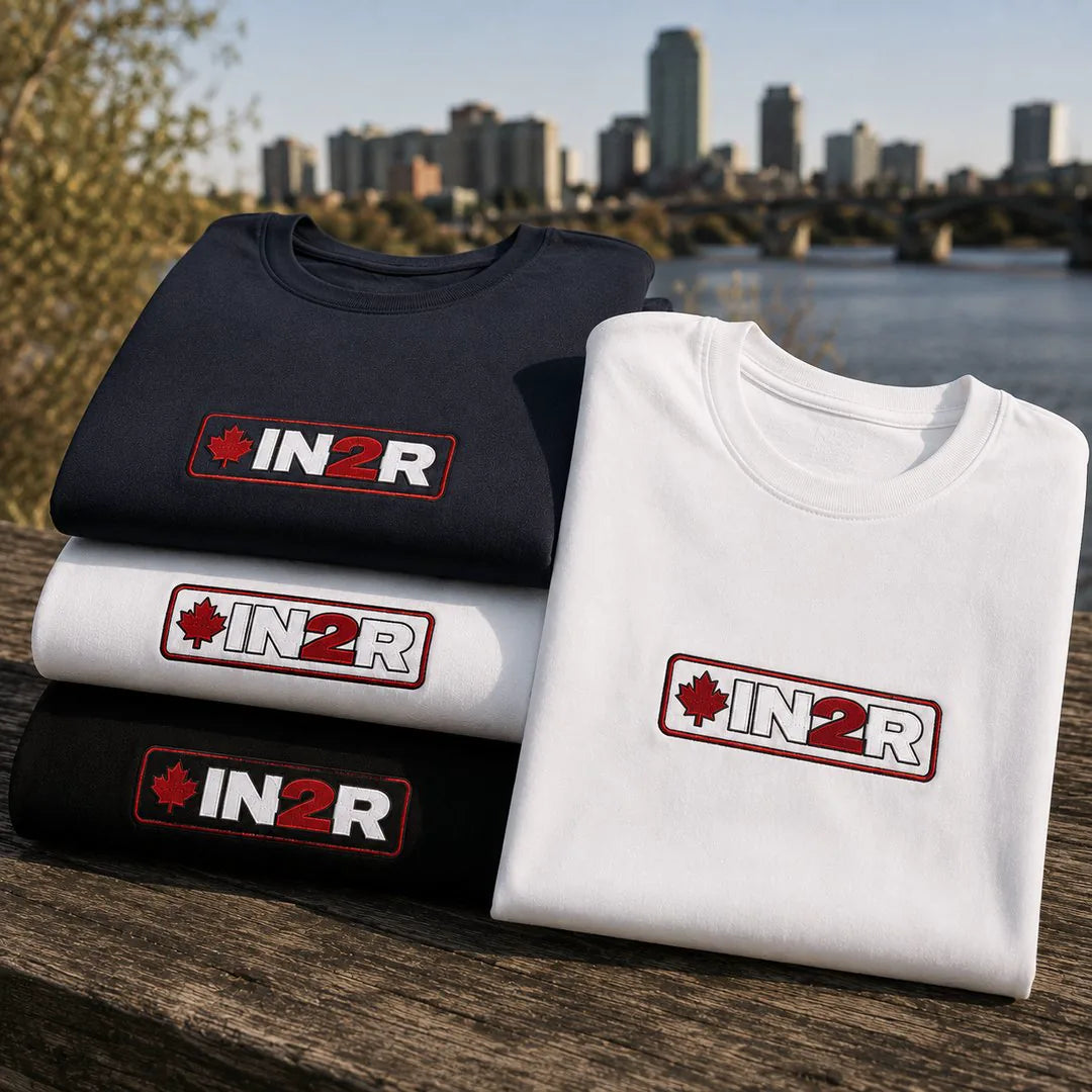 Three folded T-shirts in black, white, and black are stacked on a wooden surface outdoors, with a city skyline and river in the background. Each shirt has a rectangular chest logo reading IN2R with a red maple leaf.