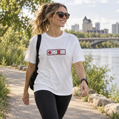A woman walking outdoors on a path by a river wearing a white graphic T-shirt with a red and black 'IN2R' logo across the chest, black leggings, sunglasses, and a black shoulder bag.