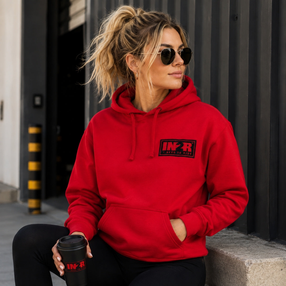 Female model wearing the IN2R Classic Red Unisex Hoodie in a modern urban setting, featuring the embroidered IN2R I Need To Ride chest patch.