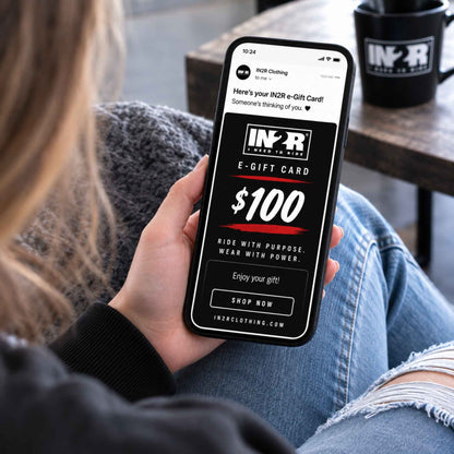 A person in a gray sweater and ripped blue jeans holds a smartphone showing an IN2R e-Gift Card $100 screen. A black IN2R mug sits on a wooden table in the background.