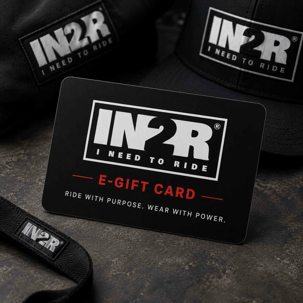 A black IN2R e-gift card with white and red text reading 'IN2R I NEED TO RIDE' and 'E-GIFT CARD', styled on a dark textured surface with black IN2R caps and a black strap in the background.