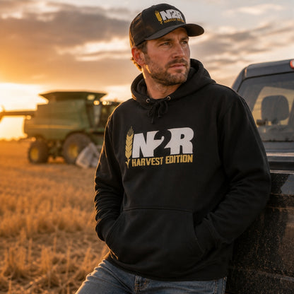 A man in a black hoodie and matching baseball cap leans against a truck in a field at sunset, with a combine harvester blurred in the background. The hoodie and cap feature a white and gold 'IN2R Harvest Edition' logo.