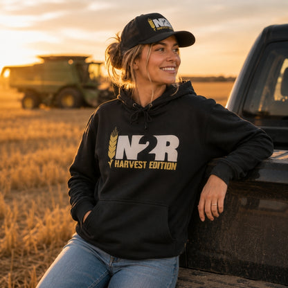 A woman wearing a black hoodie with a large white and gold 'IN2R HARVEST EDITION' graphic and a matching black baseball cap, sitting on the tailgate of a truck in a harvested field at sunset with a combine visible in the background.