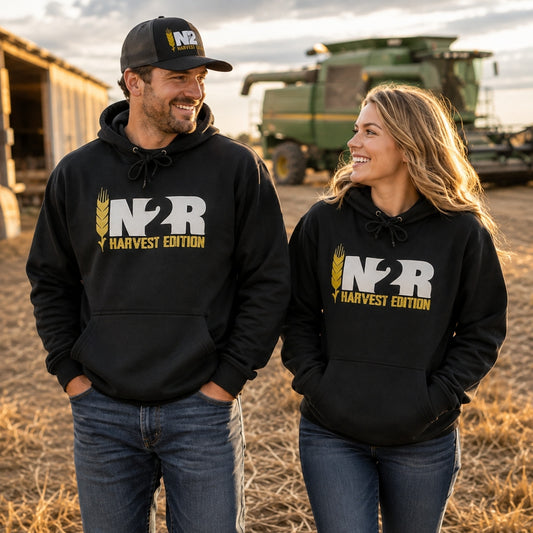 A man and a woman standing in a field wearing matching black pullover hoodies with a large white and gold IN2R Harvest Edition' graphic on the front. The man also wears a black baseball cap with the same 'IN2R' logo.
