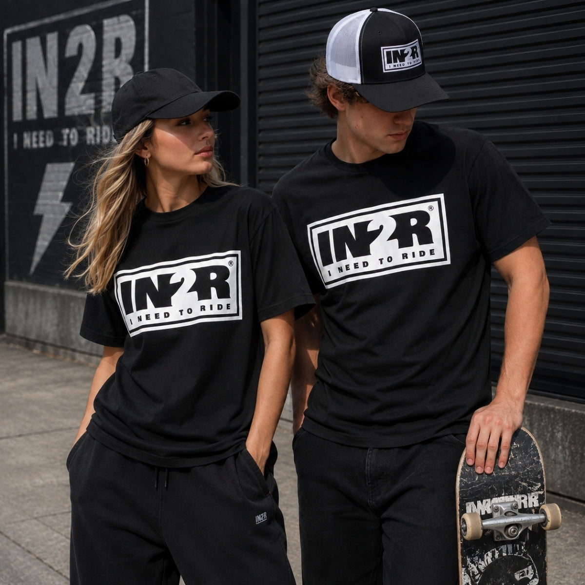 A man and a woman wearing matching black t-shirts with a white 'IN2R I NEED TO RIDE' graphic on the chest, standing outdoors in front of a dark metal shutter. The man also wears a black and white trucker hat and holds a skateboard.