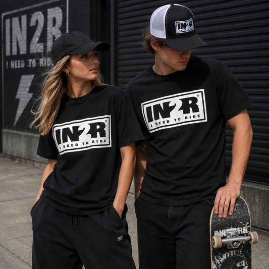 A man and a woman wearing matching black t-shirts with a white 'IN2R I NEED TO RIDE' graphic on the chest, standing outdoors in front of a dark metal shutter. The man also wears a black and white trucker hat and holds a skateboard.