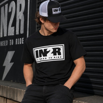 A man wearing a black t-shirt and matching trucker hat with a white and black 'IN2R I NEED TO RIDE' graphic, posed in front of a dark corrugated metal shutter.