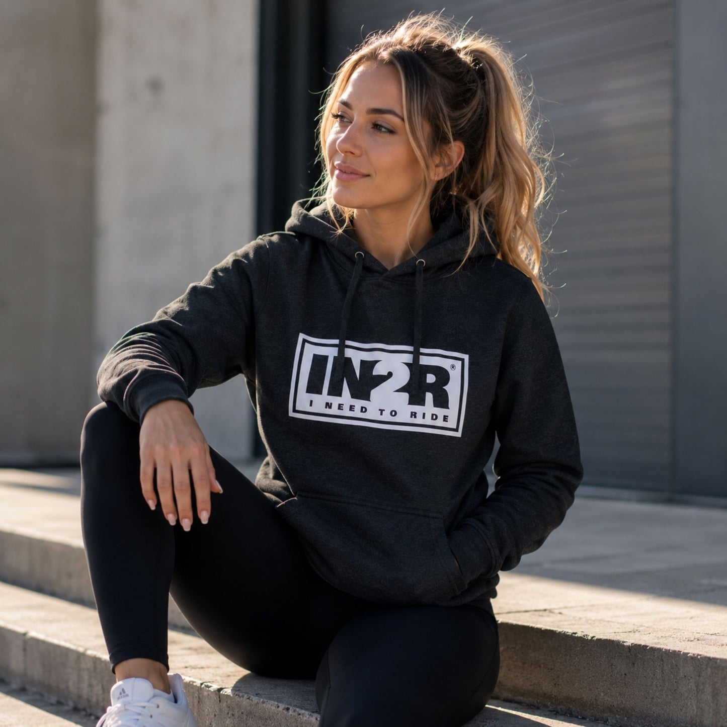 A woman wearing a dark charcoal gray hoodie with a white and black 'IN2R I NEED TO RIDE' graphic sits on concrete steps outdoors.