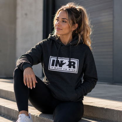 A woman wearing a dark charcoal gray hoodie with a white and black 'IN2R I NEED TO RIDE' graphic sits on concrete steps outdoors.