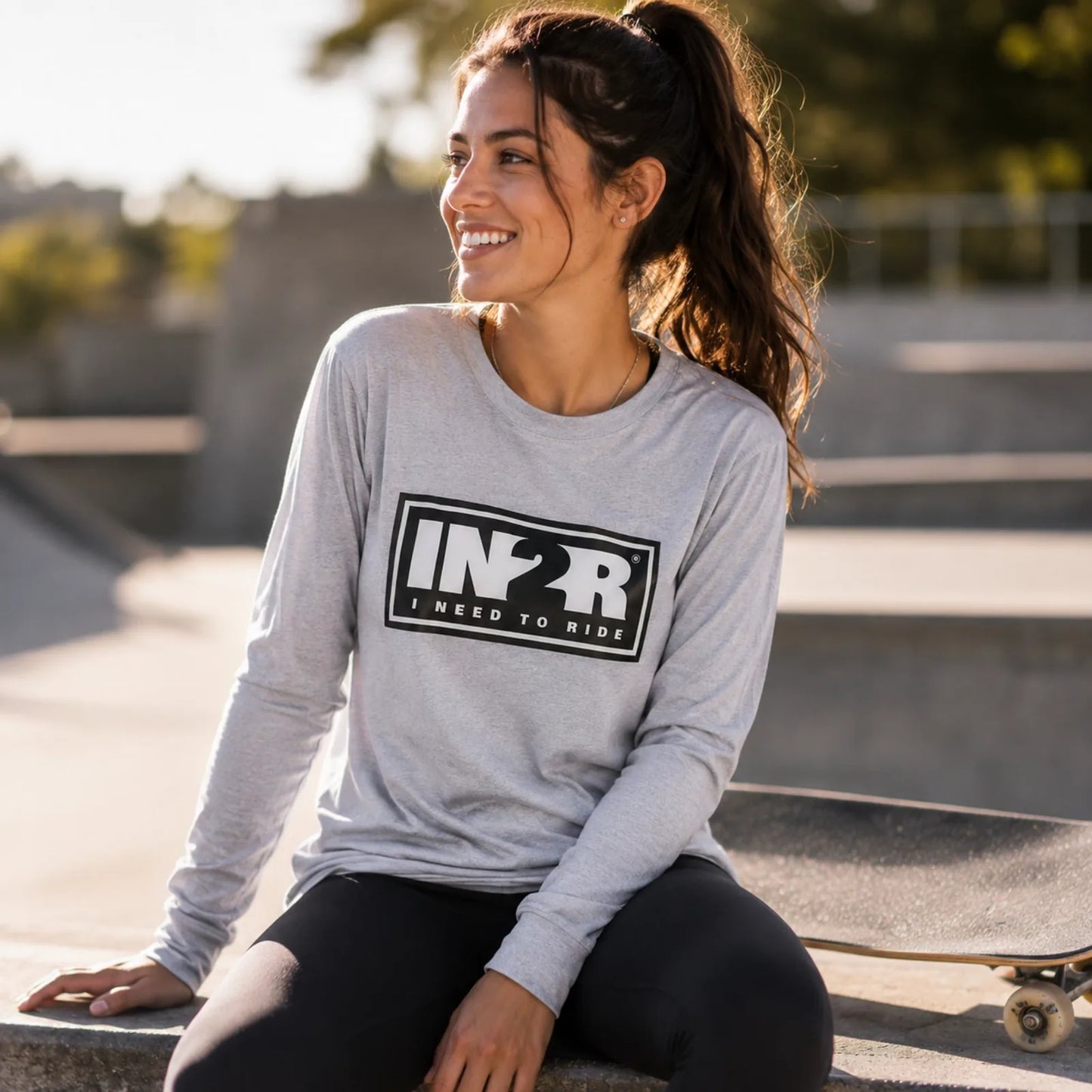A woman sitting outdoors at a skatepark wearing a light gray long-sleeve t-shirt with a black 'IN2R I NEED TO RIDE' graphic on the front, paired with black leggings.