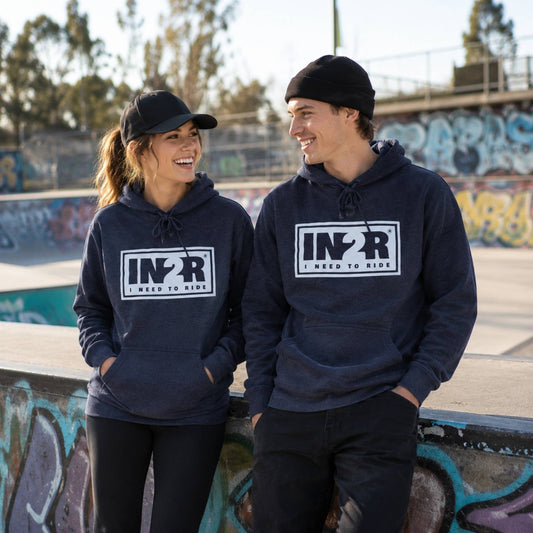 A man and a woman standing in a graffiti-covered outdoor skatepark, both wearing matching navy blue hoodies with a white 'IN2R' logo on the chest.