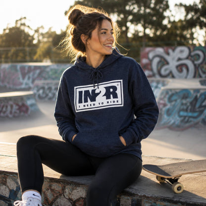 A woman sits on a concrete ledge at a skatepark wearing a navy blue hoodie with a white and black 'I Need To Ride' graphic on the chest, paired with black leggings and white sneakers, with a skateboard resting beside her.