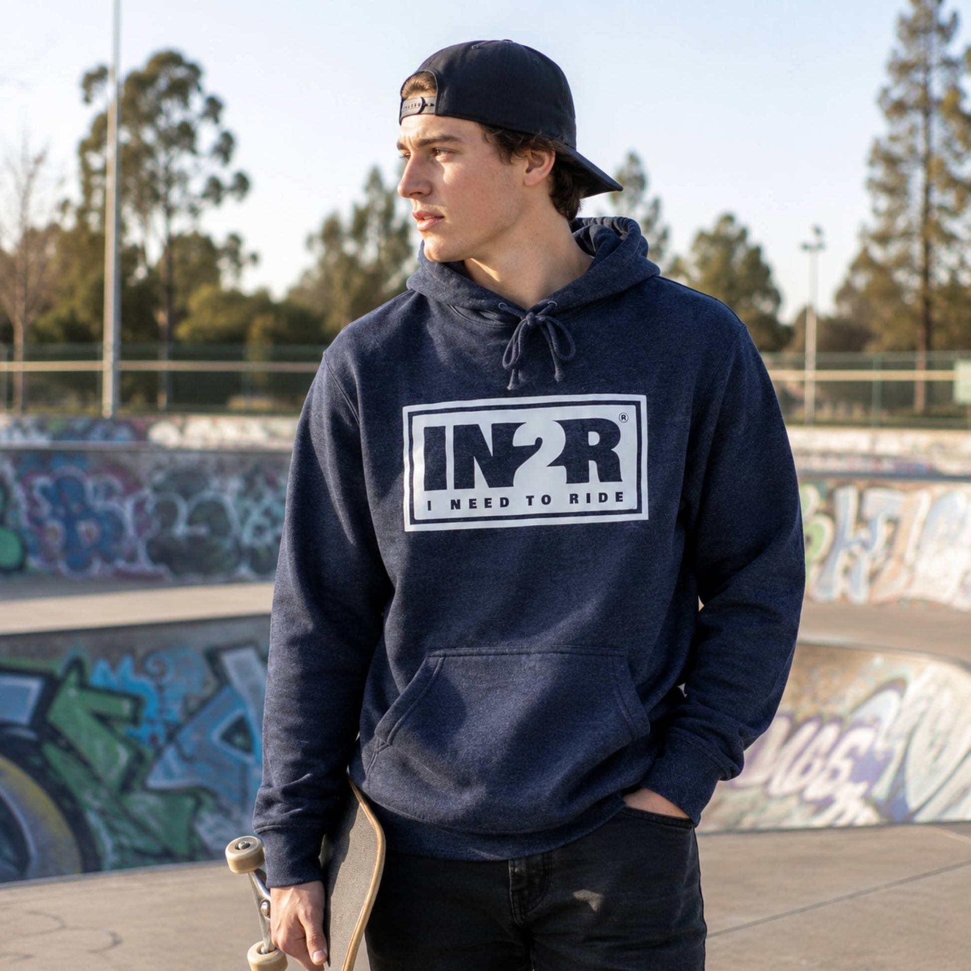 A man wearing a navy blue pullover hoodie with a white and black 'IN2R I NEED TO RIDE' graphic on the chest, standing in a graffiti-covered outdoor skatepark.