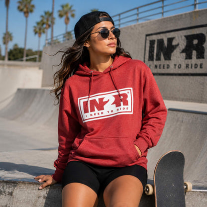 A woman wearing a red pullover hoodie with a white and red 'IN2R' logo sits on a concrete ramp at a skatepark. She is wearing a black beanie, sunglasses, and black shorts, with a skateboard resting next to her.