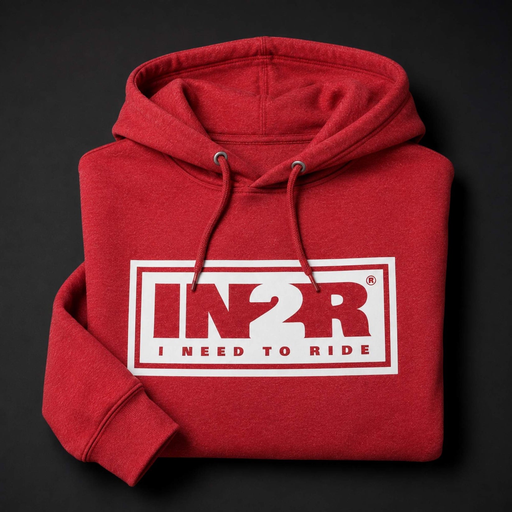A folded red hoodie with a white rectangular chest graphic reading 'IN2R I NEED TO RIDE' in red and white, shown on a dark background.