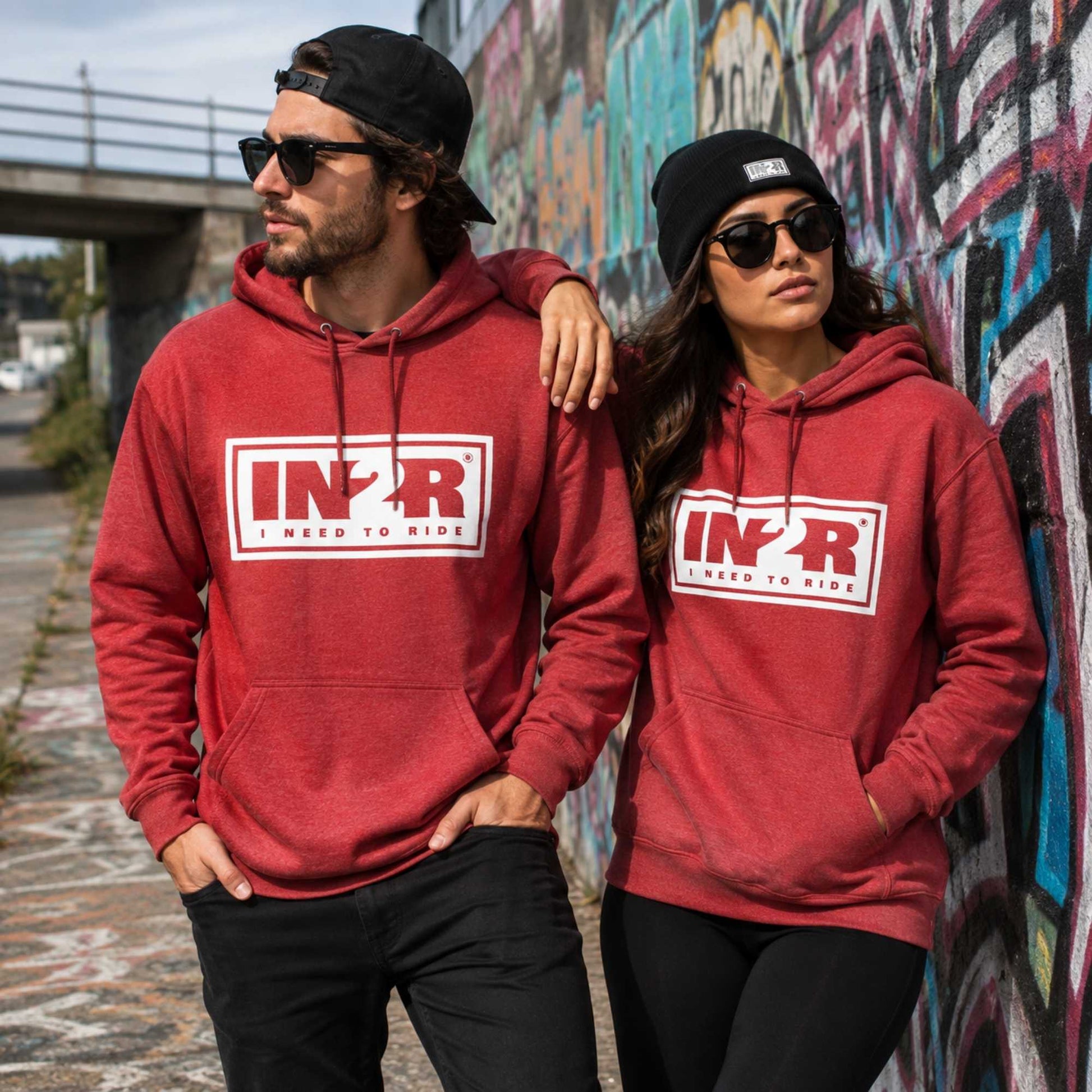 A man and a woman wearing matching red hoodies with a white and red 'IN2R' logo on the chest, standing outdoors in front of a graffiti-covered wall.