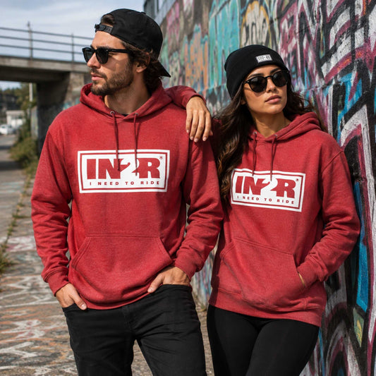 A man and a woman wearing matching red hoodies with a white and red 'IN2R' logo on the chest, standing outdoors in front of a graffiti-covered wall.