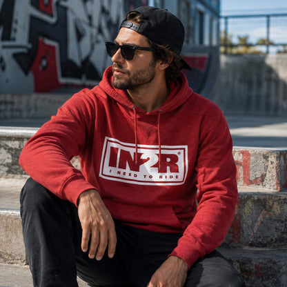 A man wearing a red pullover hoodie with a white and red 'IN2R I NEED TO RIDE' graphic on the chest, sitting on concrete steps in an outdoor urban setting.