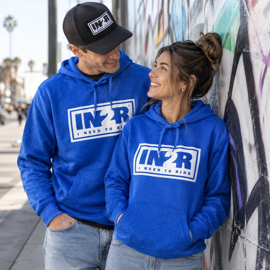 A man and a woman wearing matching royal blue hoodies with a white 'IN2R I NEED TO RIDE' graphic, standing outdoors against a graffiti-covered wall.