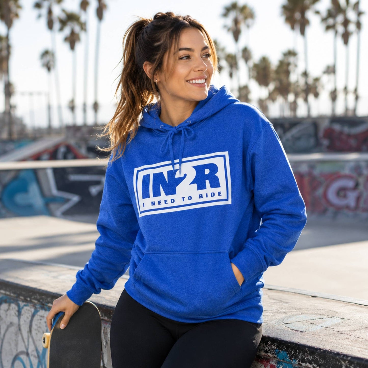 A woman wearing a bright blue pullover hoodie with a white and navy 'IN2R I Need to Ride' graphic on the chest, posing at a sunny outdoor skatepark with palm trees in the background.