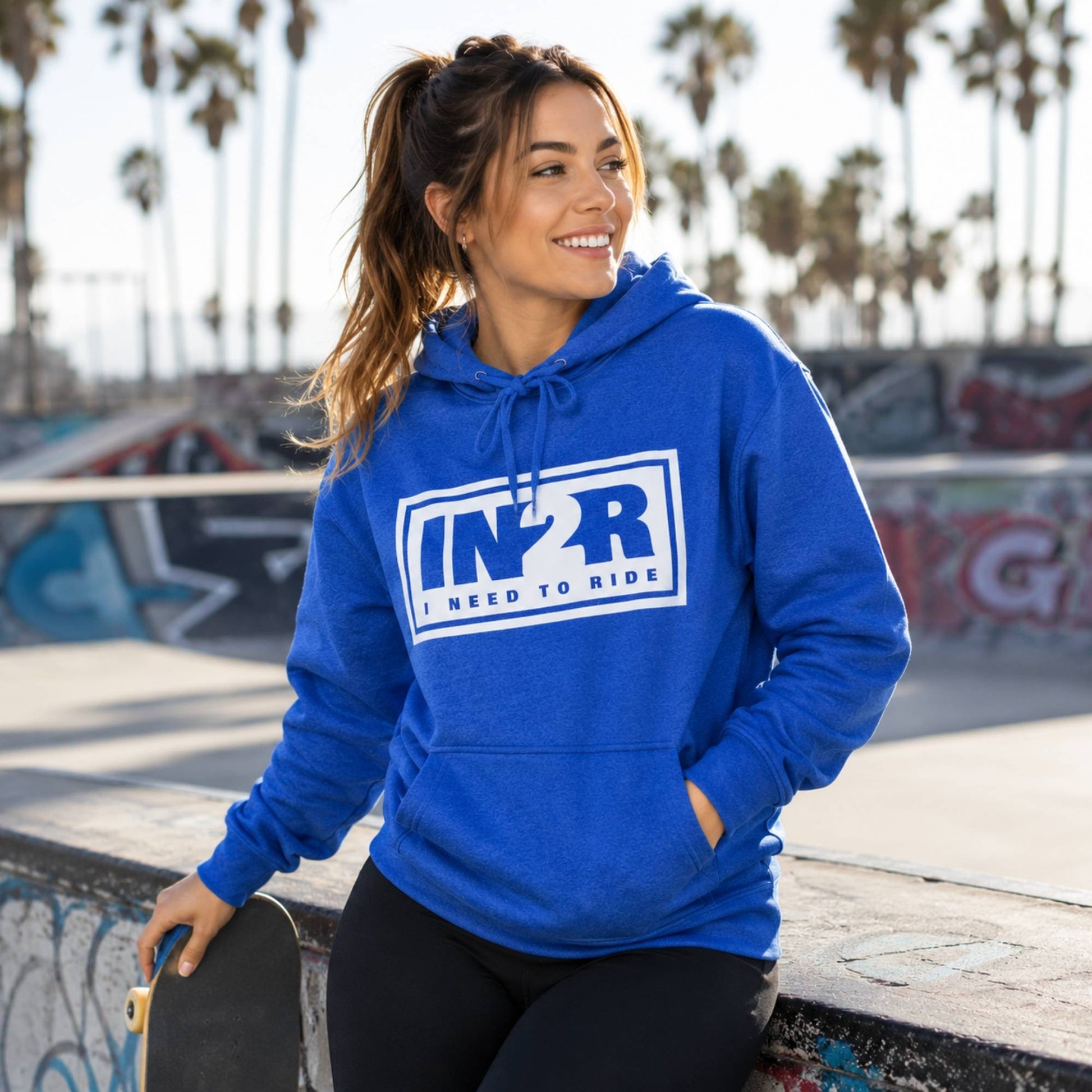 A woman wearing a bright blue pullover hoodie with a white and navy 'IN2R I Need to Ride' graphic on the chest, posing at a sunny outdoor skatepark with palm trees in the background.
