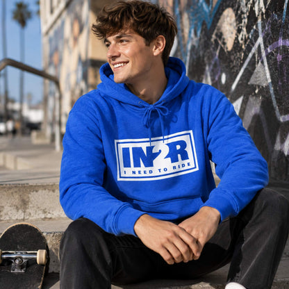 A smiling young man wearing a bright blue pullover hoodie with a white graphic that reads 'IN2R I NEED TO RIDE' sits on concrete steps outdoors, with a skateboard in the foreground and a graffiti-covered wall behind him.