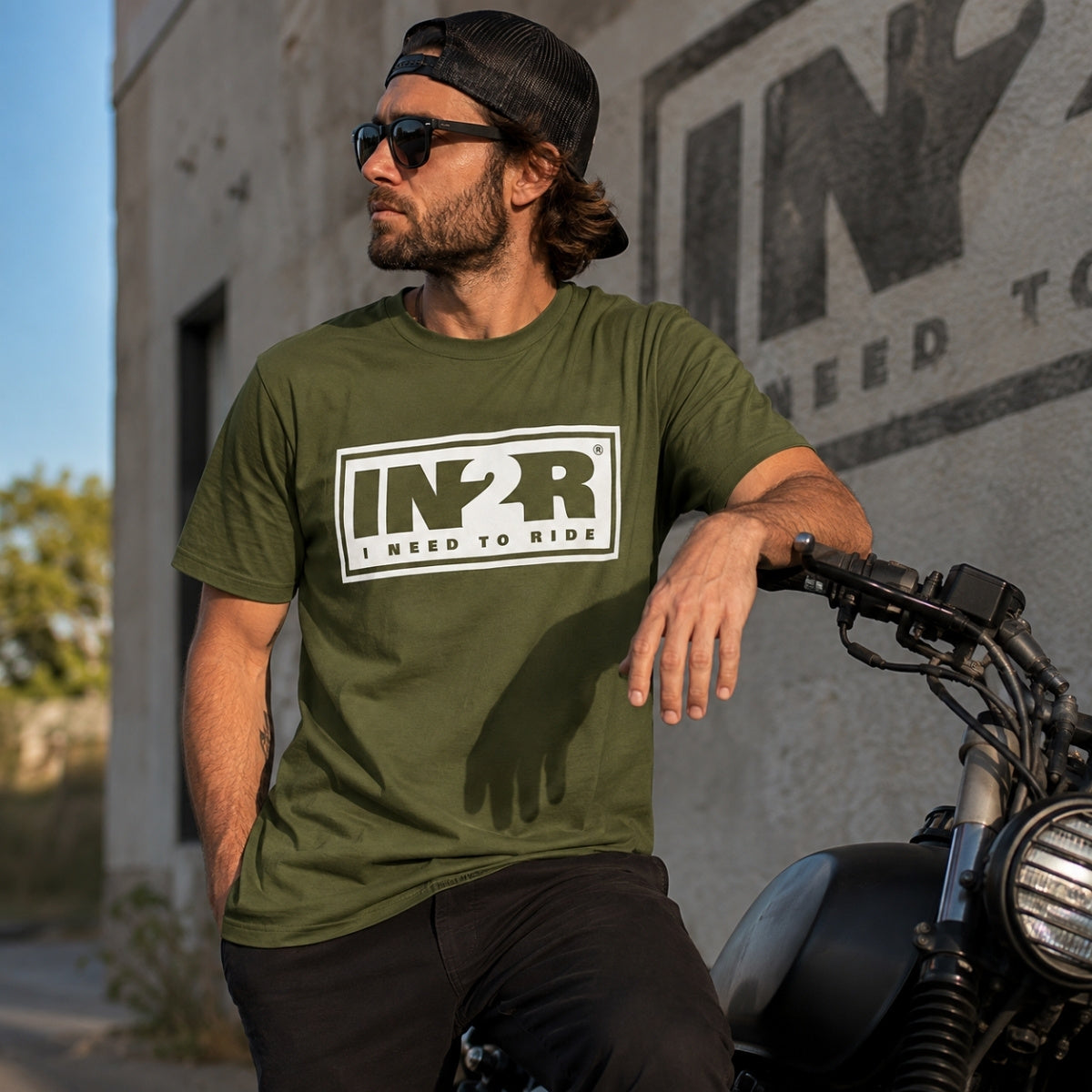 IN2R Original Moss Green Unisex T-Shirt worn by a model beside a motorcycle, showcasing the premium olive green graphic tee with oversized white IN2R logo in an urban lifestyle setting.