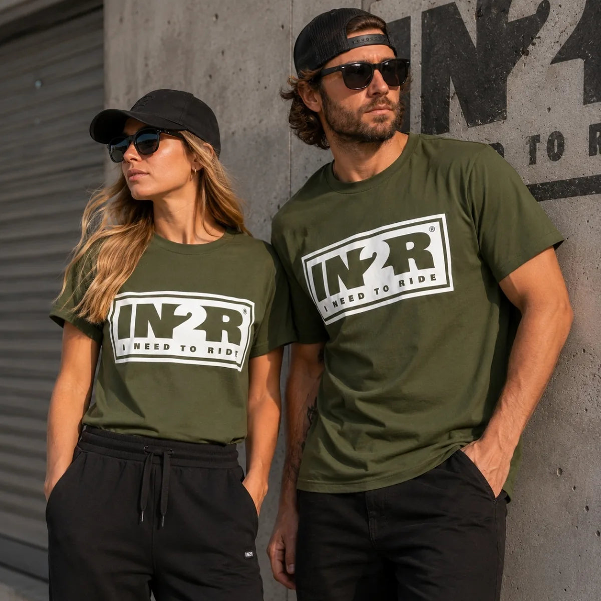 A man and a woman wearing matching olive green t-shirts with a large white and black 'IN2R I Need to Ride' logo on the chest, posing against a concrete wall.