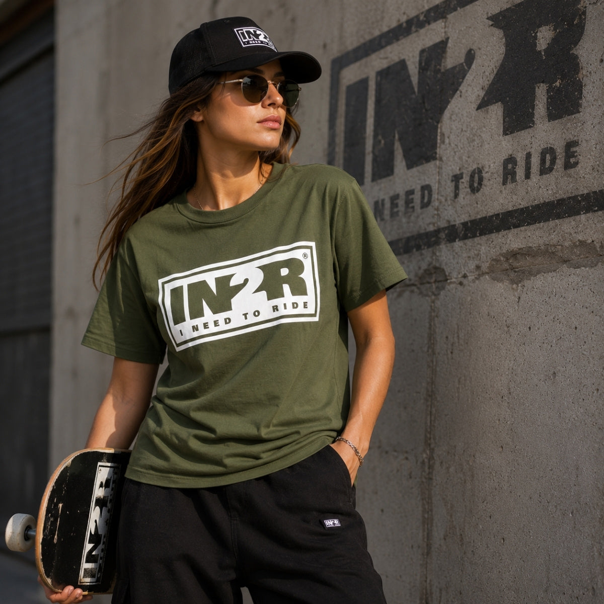 A woman wearing an olive green t-shirt with a large white and black 'IN2R NEED TO RIDE' graphic, black sweatpants, and a black cap, holding a skateboard against a concrete wall with 'IN2R NEED TO RIDE' graffiti.