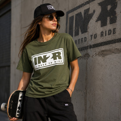 A woman wearing an olive green t-shirt with a large white and black 'IN2R NEED TO RIDE' graphic, black sweatpants, and a black cap, holding a skateboard against a concrete wall with 'IN2R NEED TO RIDE' graffiti.