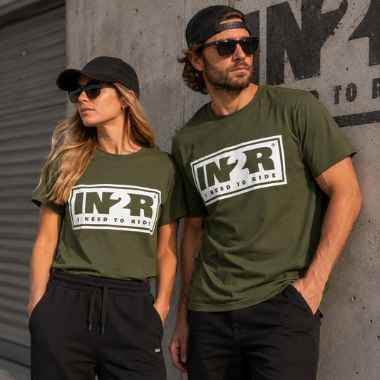 A man and a woman wearing matching moss green t-shirts with a large white and black 'IN2R I Need to Ride' logo on the chest, posing against a concrete wall.
