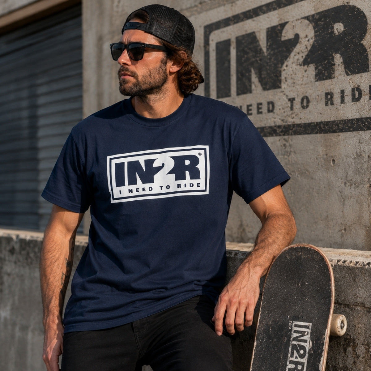 A man wearing a navy blue t-shirt with a white 'IN2R I NEED TO RIDE' graphic, sunglasses, and a black cap, leaning on a skateboard against a concrete wall with a matching IN2R logo.