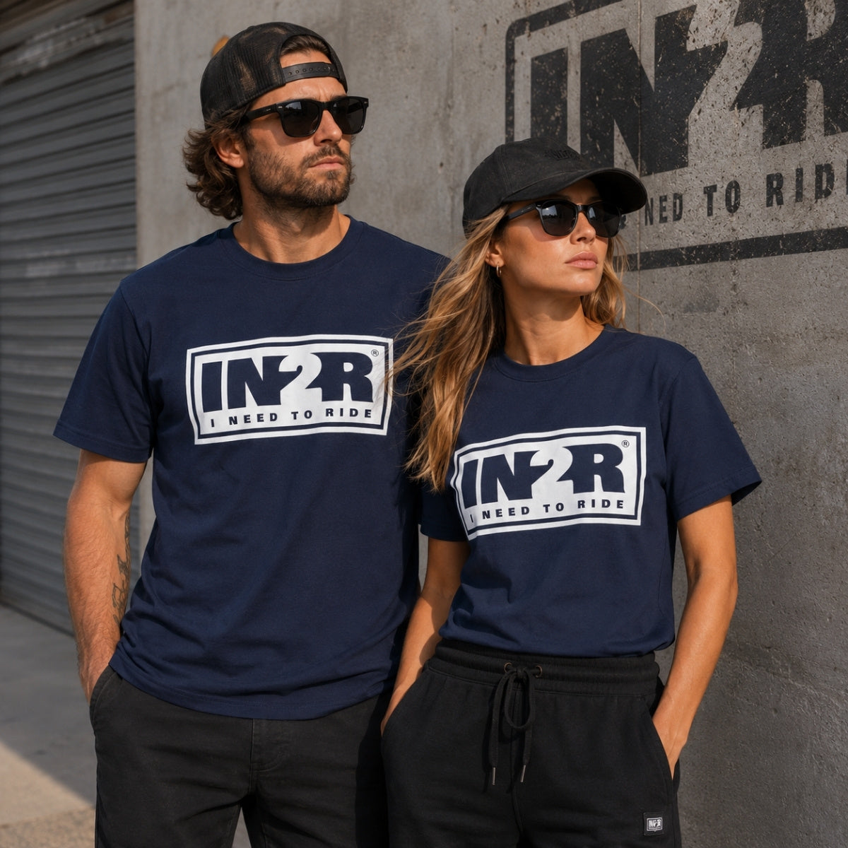 A man and a woman wearing matching navy blue t-shirts with a white and black 'IN2R I NEED TO RIDE' graphic on the chest, standing against a concrete wall with a large IN2R logo.