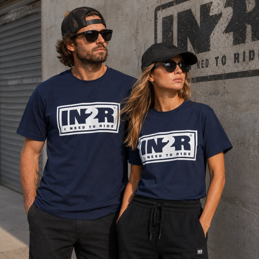 A man and a woman wearing matching navy blue t-shirts with a white and black 'IN2R I NEED TO RIDE' graphic on the chest, standing against a concrete wall with a large IN2R logo.