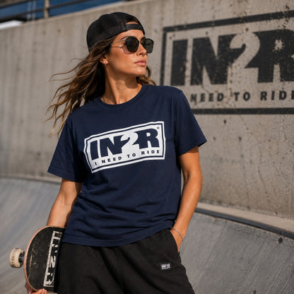 A woman wearing a navy blue t-shirt with a white and black 'I Need To Ride' logo, black sweatpants, a black cap, and sunglasses, holding a skateboard in a concrete skatepark.