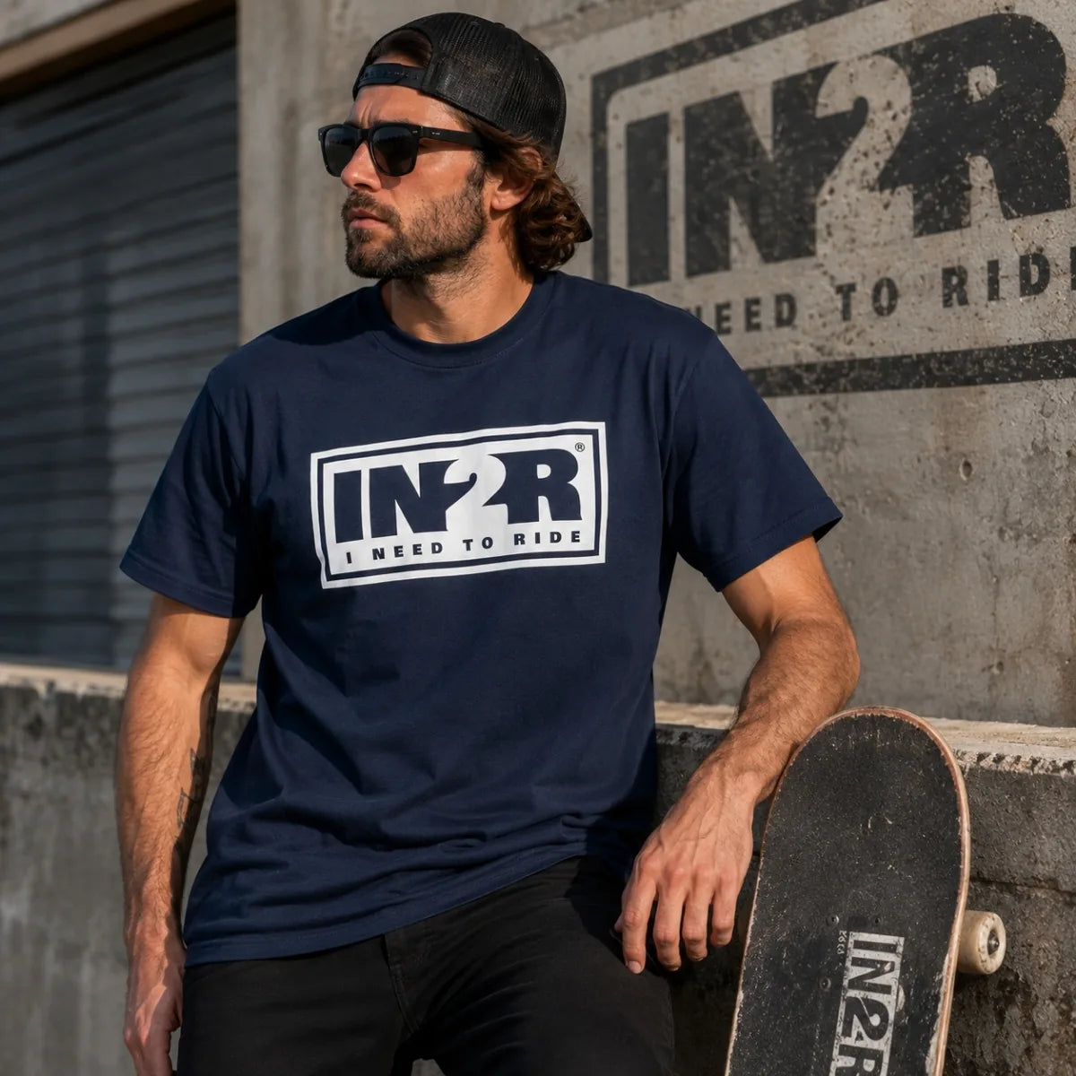 A man wearing a navy blue t-shirt with a white 'IN2R I NEED TO RIDE' graphic, sunglasses, and a black cap, leaning on a skateboard against a concrete wall with a matching IN2R logo.
