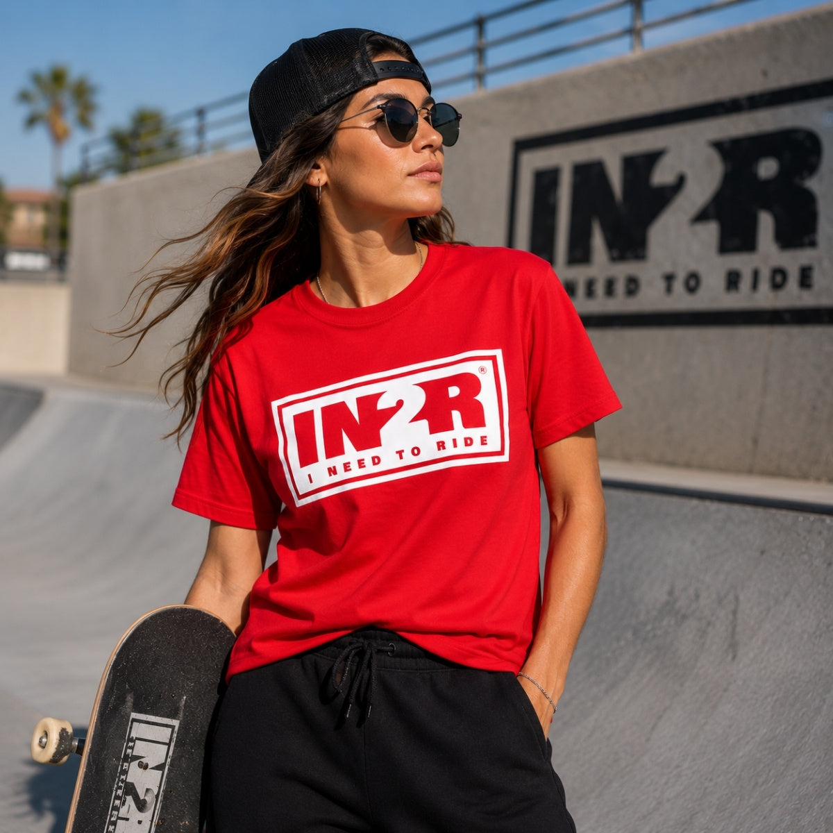 A woman wearing a red t-shirt with a white 'IN2R I NEED TO RIDE' graphic, black sweatpants, and a black cap, standing at a skatepark holding a skateboard.