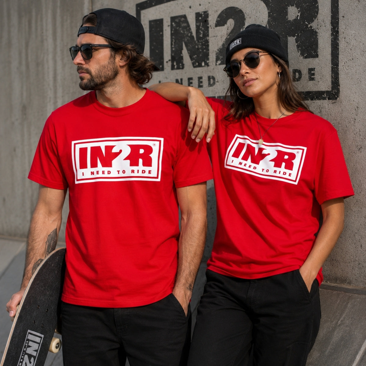 A man and a woman wearing matching bright red t-shirts with a white and black 'IN2R I NEED TO RIDE' logo on the chest, posing together against a concrete wall.