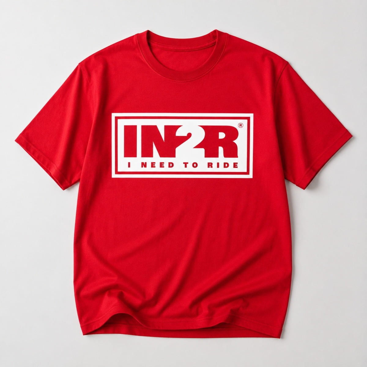 A red short-sleeve crewneck t-shirt with a white graphic print on the chest reading 'IN2R I NEED TO RIDE' inside a white rectangular border, shown on a light gray background.