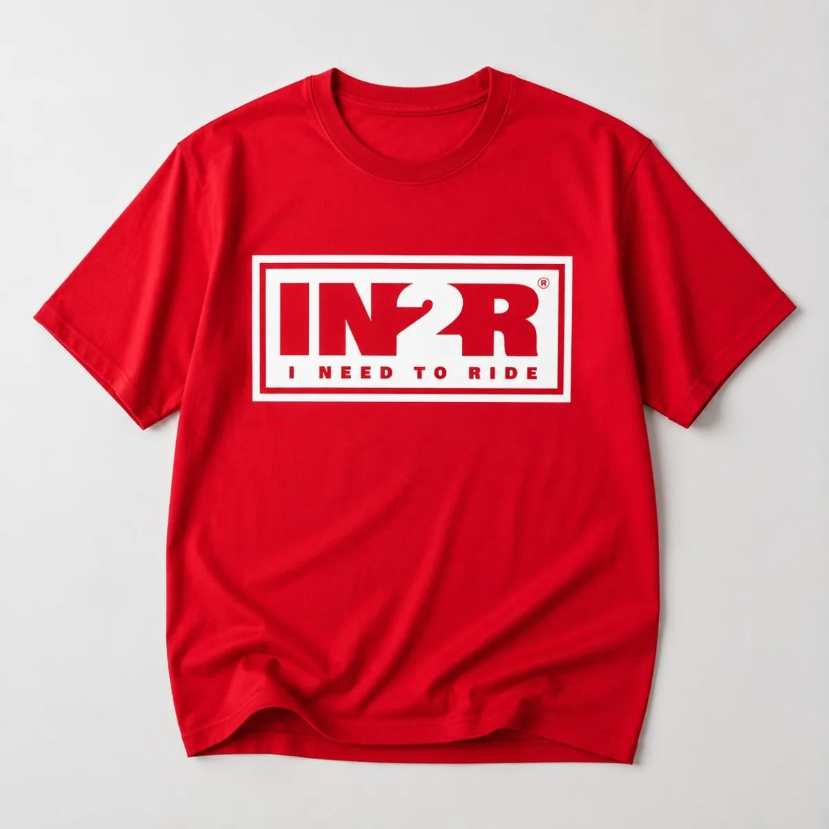 A red short-sleeve crewneck t-shirt with a white graphic print on the chest reading 'IN2R I NEED TO RIDE' inside a white rectangular border, shown on a light gray background.
