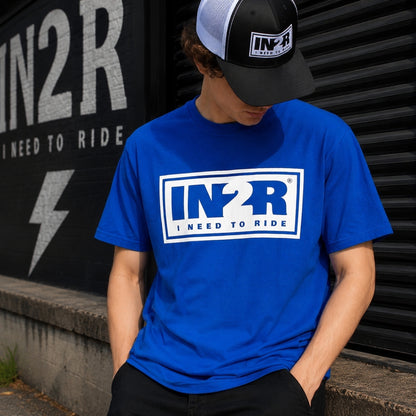 A person wearing a bright blue short-sleeve t-shirt with a white and blue 'IN2R I NEED TO RIDE' logo on the chest, paired with a black and white trucker hat. The person is leaning against a black corrugated metal wall with a matching 'IN2R' sign in the background.