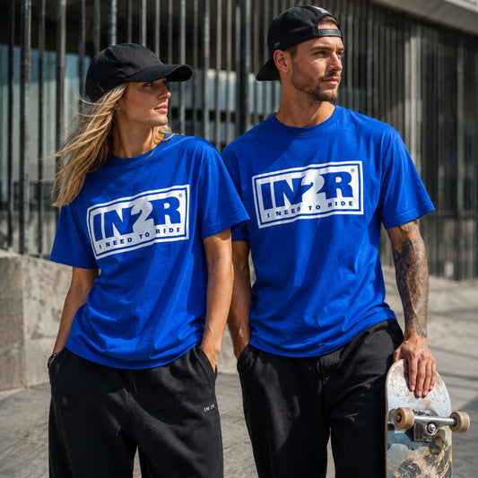 A man and a woman wearing matching royal blue t-shirts with a white 'IN2R I NEED TO RIDE' logo on the chest, standing outdoors in front of a metal fence. The man is holding a skateboard.