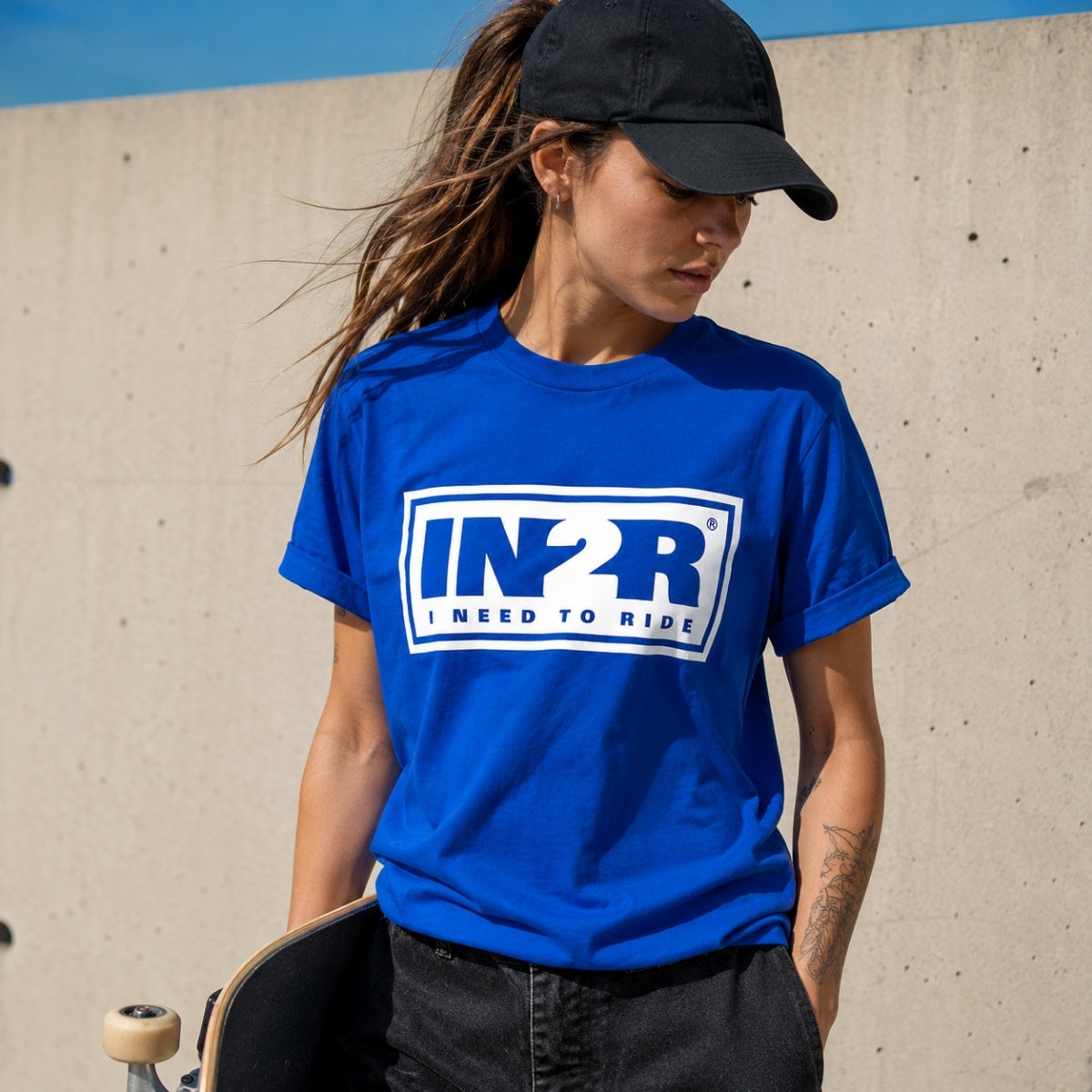 A woman wearing a bright blue graphic t-shirt with a white and blue 'IN2R I NEED TO RIDE' logo, paired with a black baseball cap and dark jeans, holding a skateboard against a concrete wall.