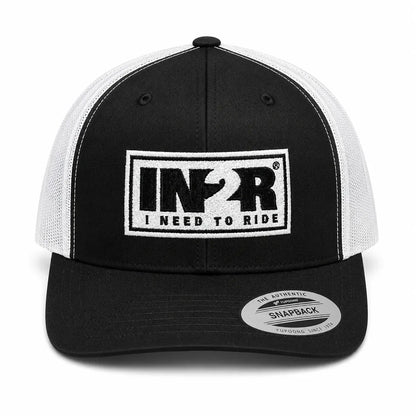 A black and white trucker hat with a black front panel, white mesh back, and a white rectangular patch on the front that reads 'IN2R RIDE I NEED TO RIDE'. A silver 'Snapback' sticker is visible on the brim.