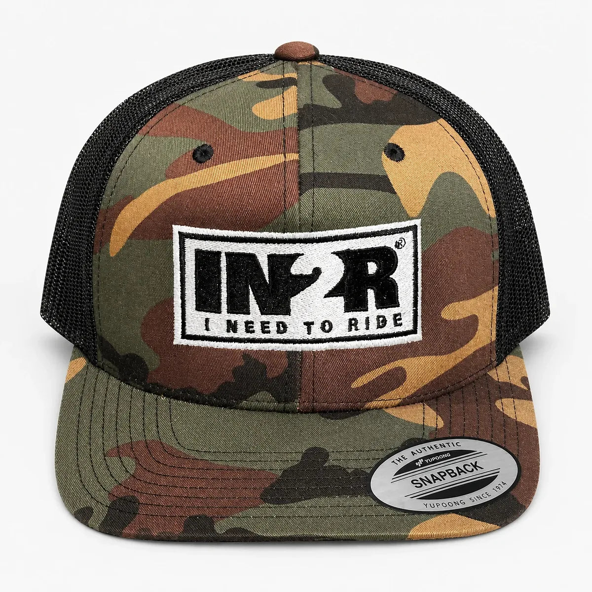 A camouflage trucker hat with a black mesh back and a front patch reading 'IN2R I NEED TO RIDE'. A circular 'The Authentic Snapback' sticker is visible on the brim.